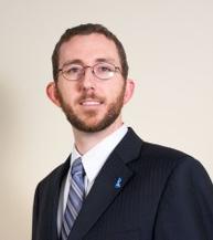 Chest up of a man with shirt brown hair, short brown beard and glasses, wearing a dark blue suit with white shirt and light blue striped tie