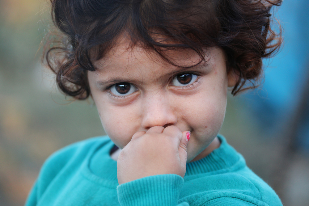 Closeup of female toddler with hand in mouth, very big brown eyes, short curly brown hair and turquoise sweatshirt