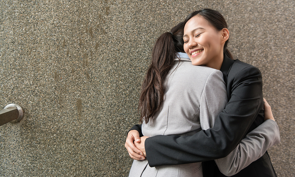 Smiling woman in black suit hugs woman in grey suit in front of concrete wall