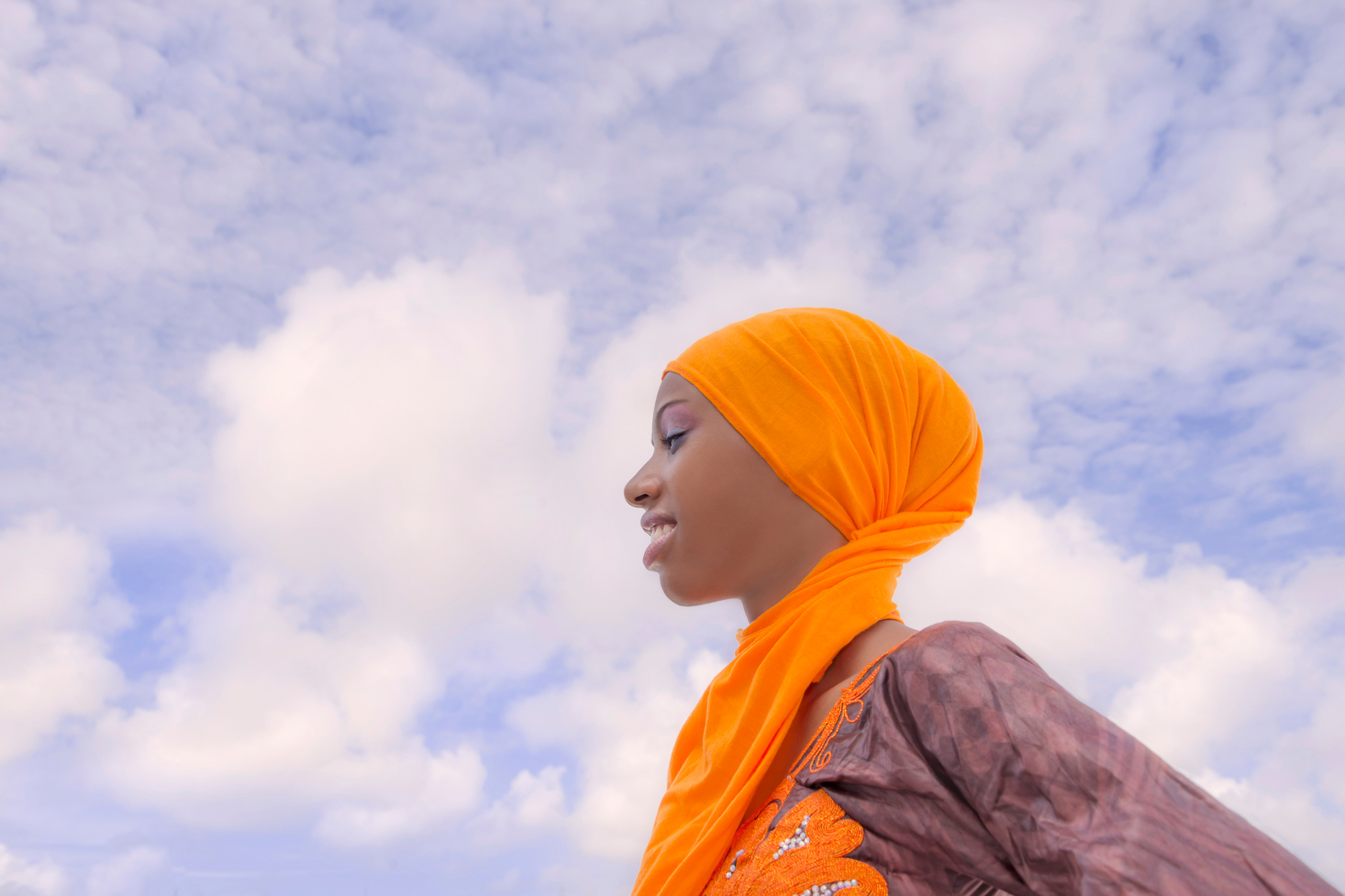Profile of a young African woman in orange head wrap set against a blue sky wit clouds